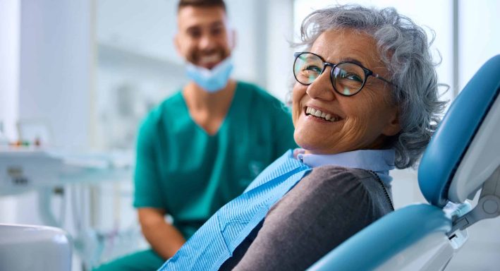 Happy senior woman at dental clinic looking at camera. Her dentist is in the background.