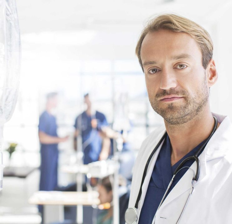 Portrait of mid adult doctor with colleagues in background, standing in hospital ward